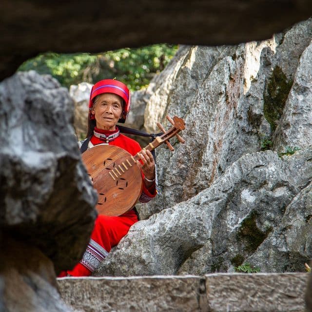 Eine ältere Frau in traditioneller roter Tracht und Kopfbedeckung sitzt zwischen Felsen und hält ein rundes hölzernes Saiteninstrument.