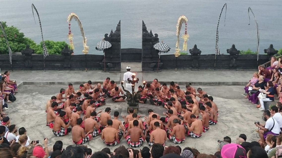 Un grupo de hombres sin camisa con pareos se sientan en círculo para una ceremonia de fuego tradicional en un escenario con vistas al mar.