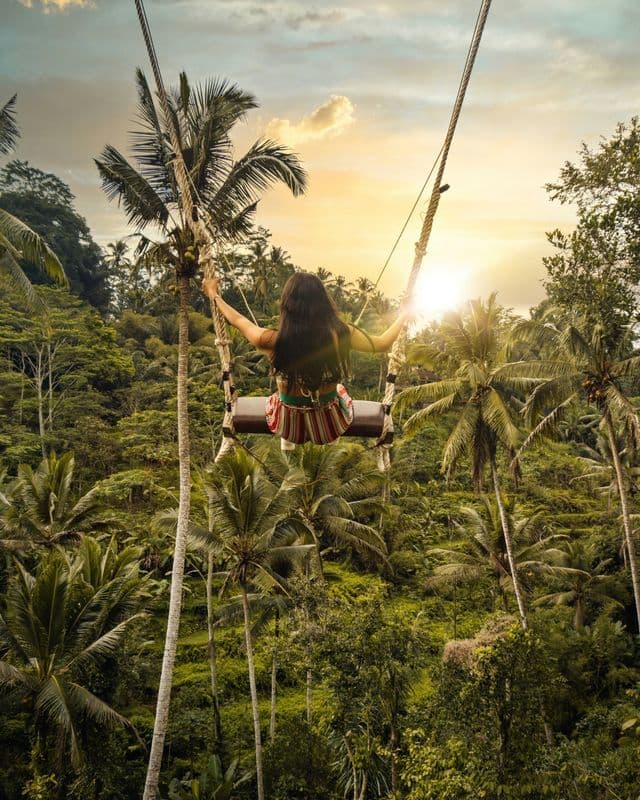 Una mujer vista de espaldas en un columpio alto, volando sobre una densa selva tropical con palmeras al atardecer.