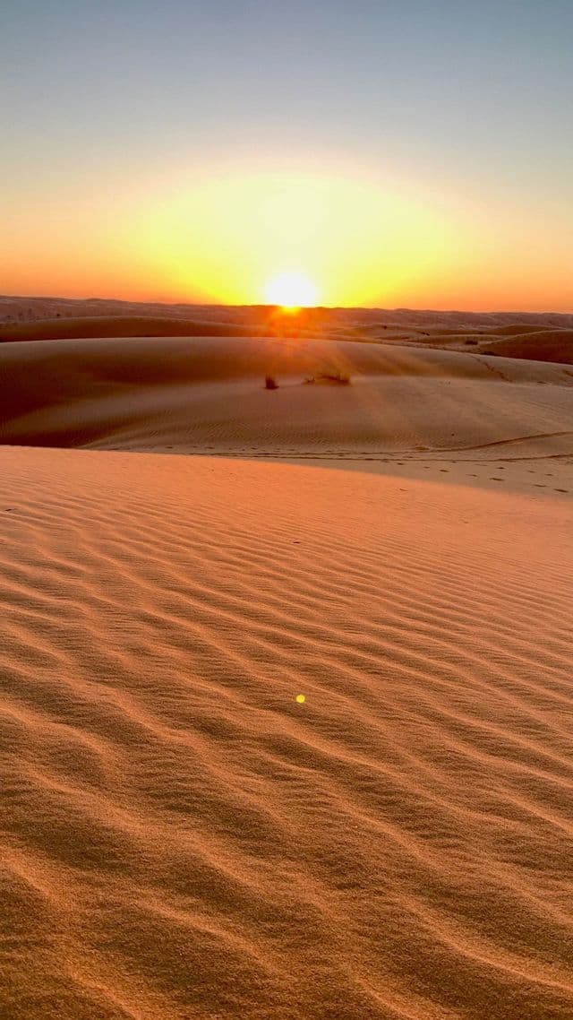 El sol se pone sobre vastas dunas de arena del desierto, proyectando una cálida luz naranja sobre la arena ondulada en primer plano.