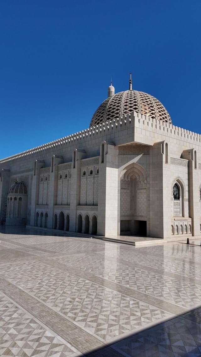 Una mezquita de piedra ornamentada con una gran cúpula estampada y arcos, vista desde su patio embaldosado bajo un cielo azul claro.