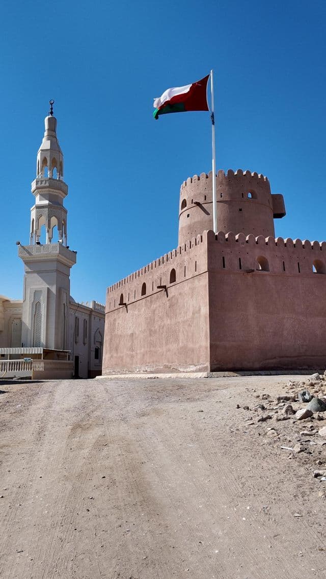 Un fuerte marrón rojizo con torre redonda se alza junto a un minarete de mezquita blanco, con la bandera de Omán ondeando bajo un cielo azul claro.