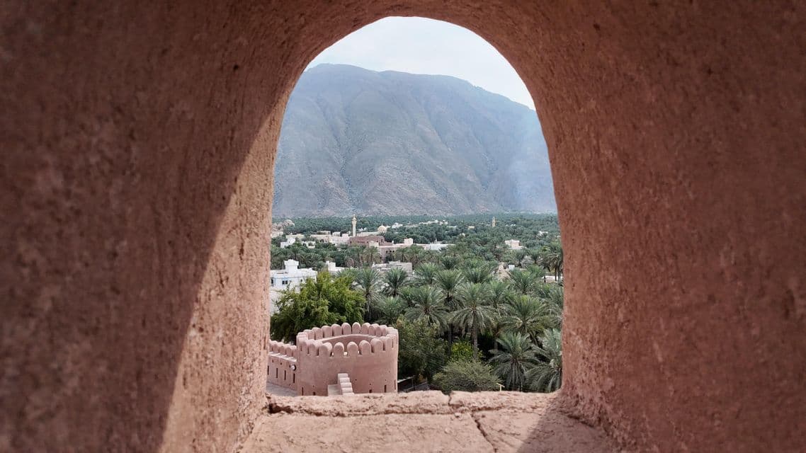 Una torre fortaleza y un oasis de palmeras vistos a través de un arco de piedra, con una montaña al fondo.