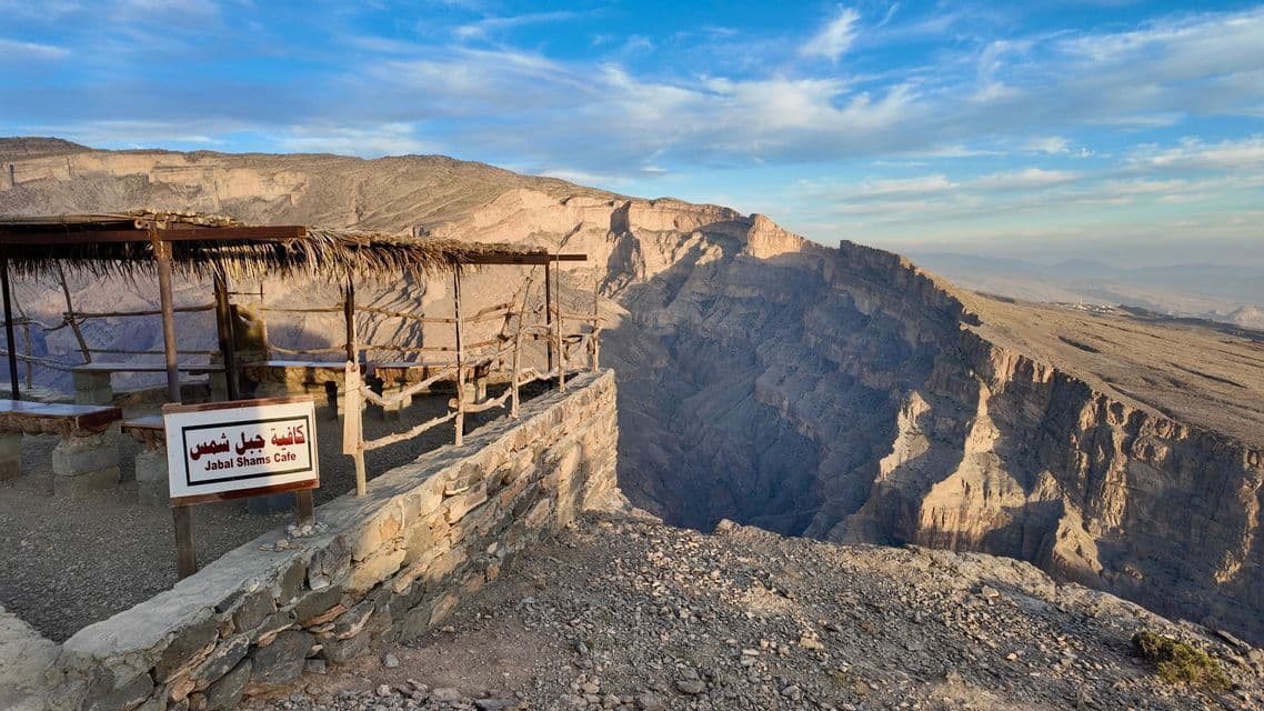 Un refugio rústico con techo de paja se alza en un mirador de piedra con vistas a un vasto cañón rocoso bajo un cielo parcialmente nublado.