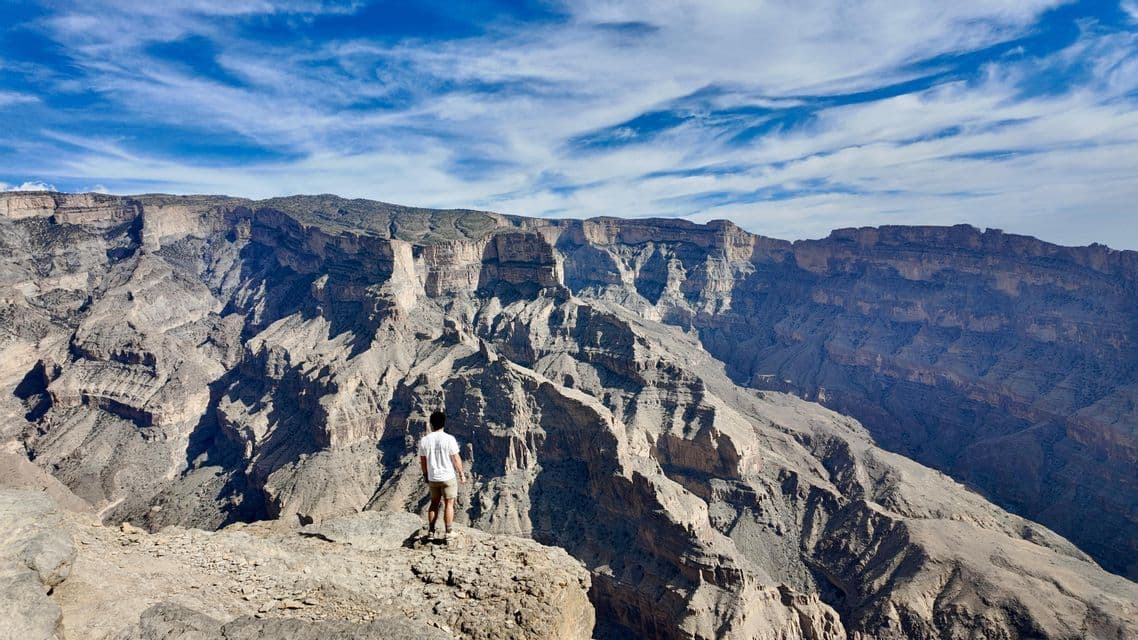 Una persona se para al borde de un acantilado, mirando un cañón profundo y rocoso bajo un cielo azul con nubes.
