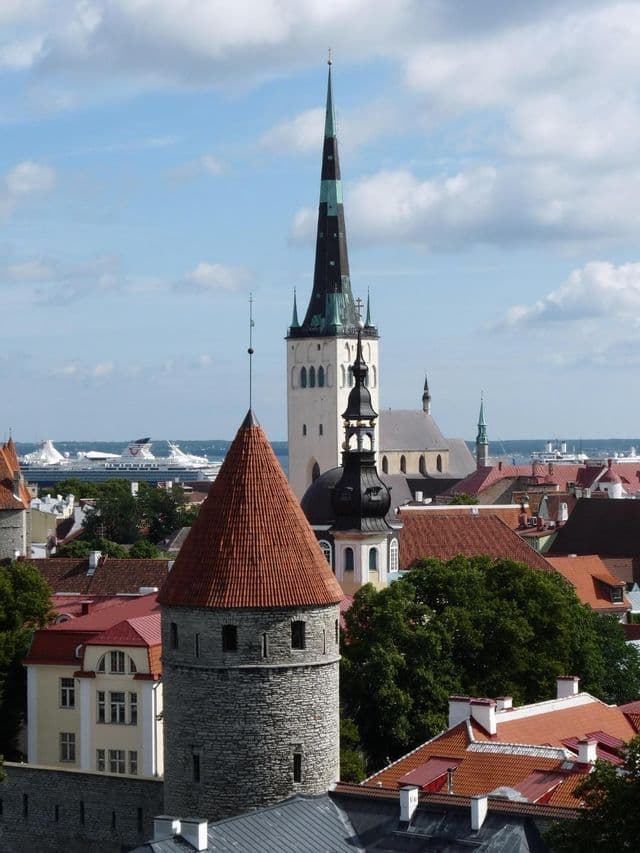 Eine Luftaufnahme einer historischen Stadt mit einem Steinturm mit rotem Dach, einem hohen Kirchturm und einem Kreuzfahrtschiff auf dem Wasser.