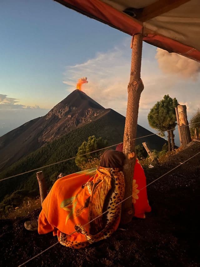 Une personne enveloppée dans une couverture orange observe depuis un camping l'éruption d'un volcan avec de la fumée et de la lave au loin.