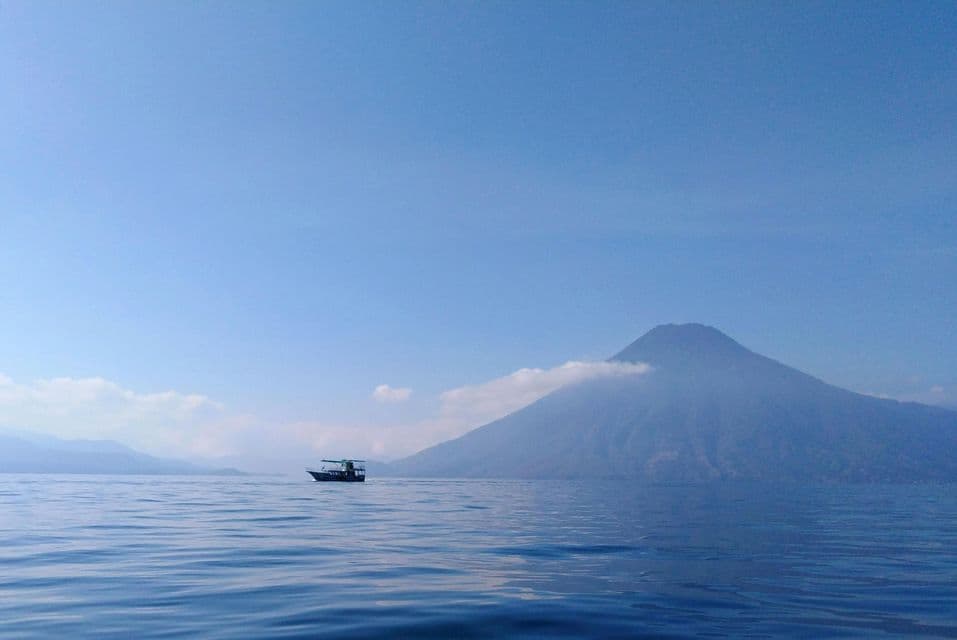 Un petit bateau navigue sur des eaux calmes devant un grand volcan partiellement couvert de nuages sous un ciel bleu clair.