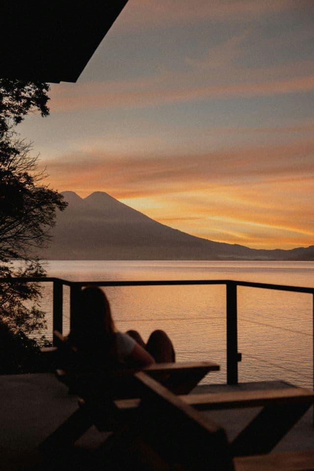 Une personne en silhouette est assise sur une chaise de balcon, regardant le coucher de soleil sur un lac tranquille et une chaîne de montagnes lointaine.