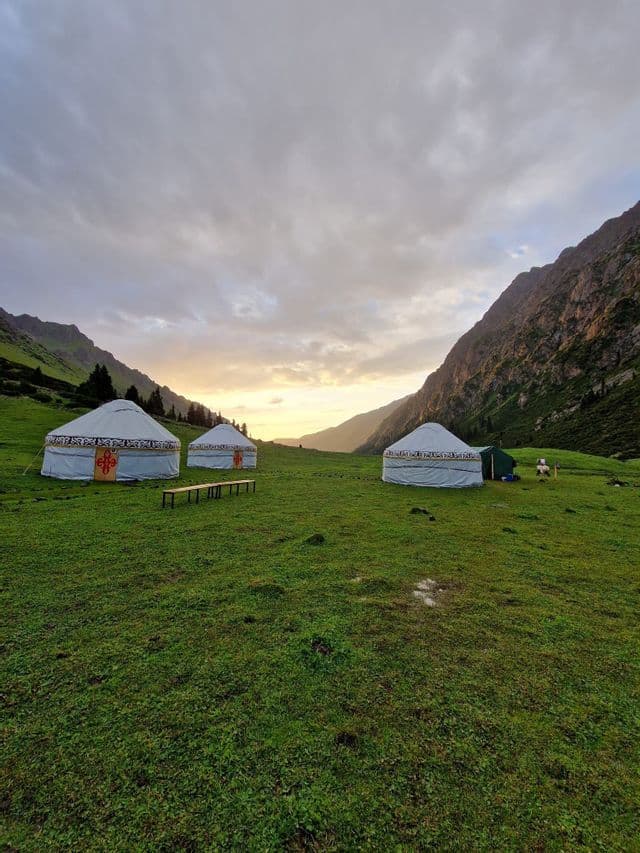 Three traditional yurts sit on a grassy field in a mountain valley during a cloudy sunset.