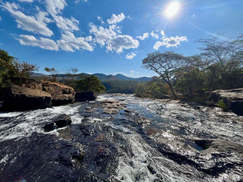 Un ampio fiume scorre a cascata su rocce scure e bagnate sotto un sole splendente e un cielo blu con nuvole bianche.