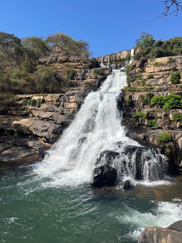 Una potente cascata scende lungo una serie di formazioni rocciose a terrazze, confluendo in una pozza verde spumeggiante sotto un cielo azzurro limpido.