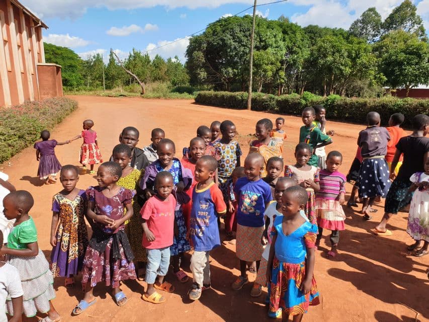 Un folto gruppo di bambini con abiti colorati si trova su una strada sterrata di fronte a un edificio e alberi.