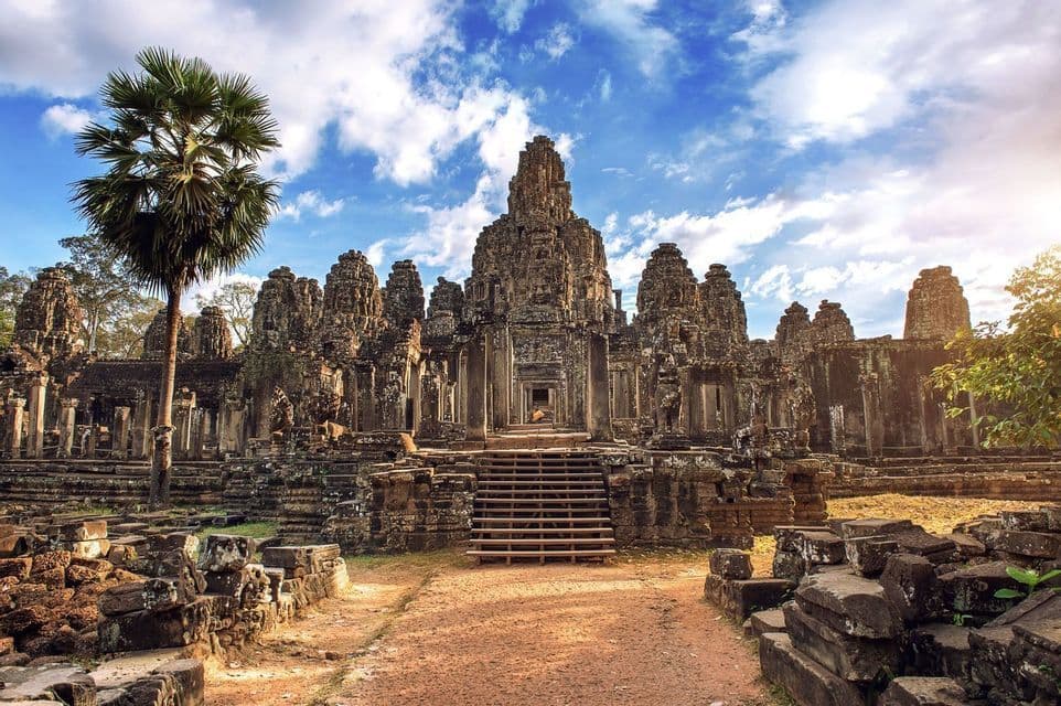 Un grand temple de pierre ancien avec des tours sculptées et un escalier central, avec un palmier à gauche sous un ciel partiellement nuageux.