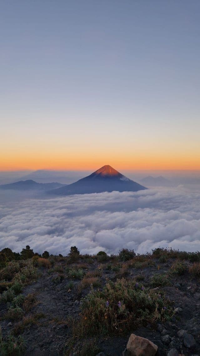 Una cima vulcanica emerge da una spessa coltre di nuvole all'alba, vista da un punto panoramico roccioso.