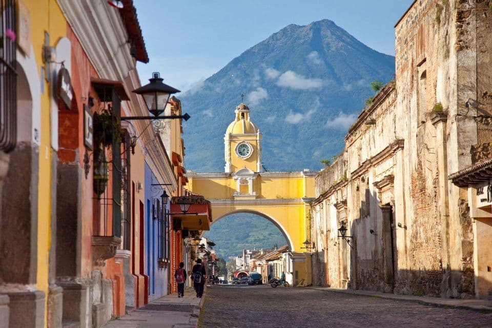 Un arco giallo con una torre dell'orologio si erge su una strada acciottolata fiancheggiata da edifici colorati, con un grande vulcano sullo sfondo.