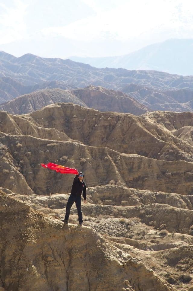 Una persona è in piedi su uno sperone roccioso, con la bandiera del Kirghizistan, e vista su canyon aridi.