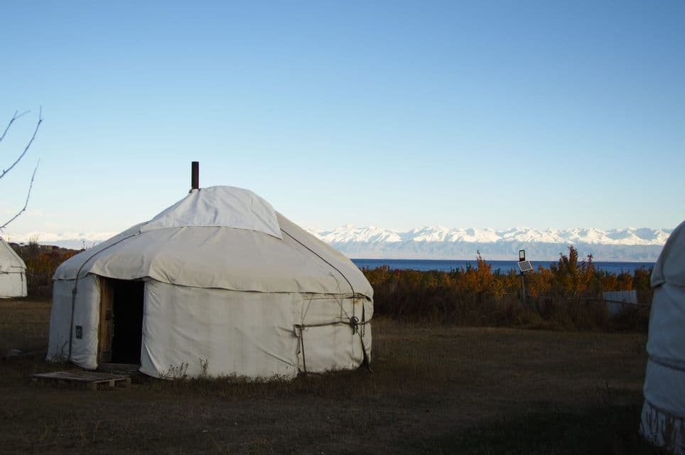 Una yurta bianca si erge in un campo con un lago e montagne innevate sullo sfondo sotto un cielo azzurro e limpido.