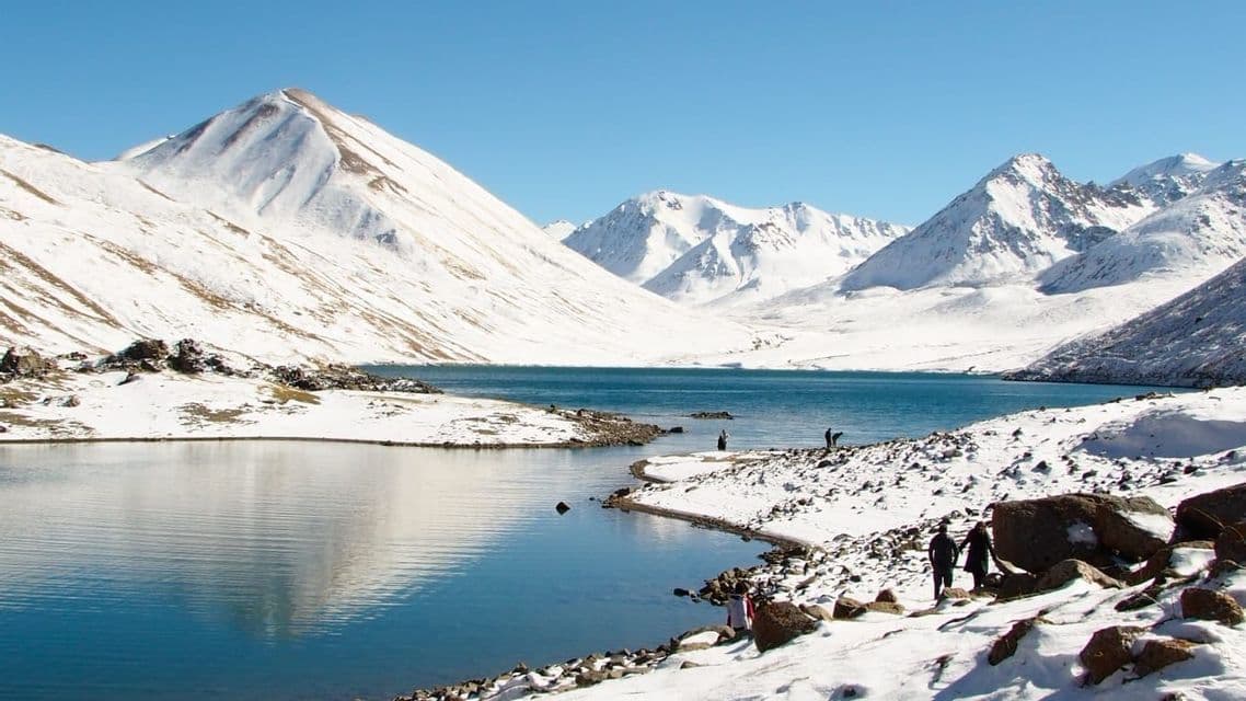 Eine WeRoad Gruppenreise spaziert am felsigen, schneebestäubten Ufer eines ruhigen Sees, umgeben von großen, schneebedeckten Bergen.