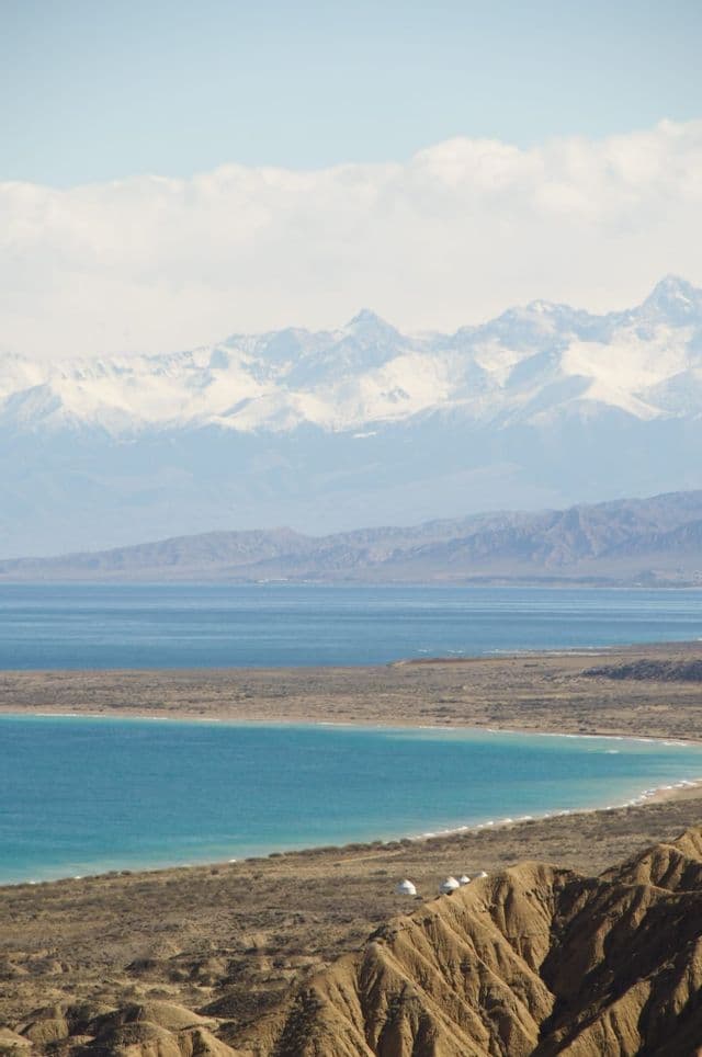 Una vista dall'alto di un lago turchese e una costa arida, con una catena montuosa innevata in lontananza sotto un cielo velato.