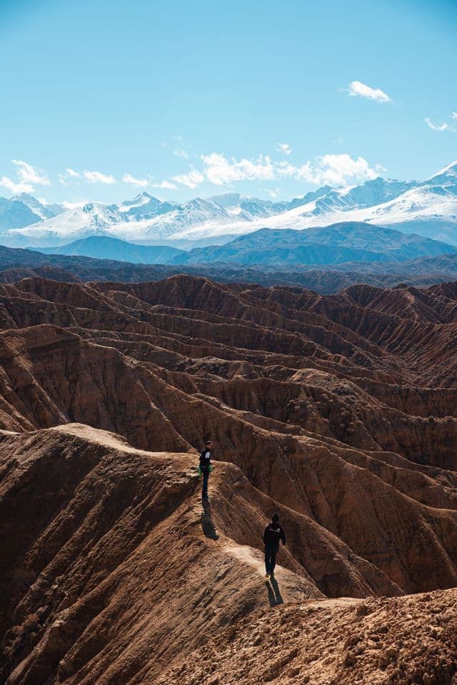 Due persone in un viaggio di gruppo WeRoad camminano lungo un sentiero sterrato su una cresta di canyon con uno sfondo di montagne innevate.