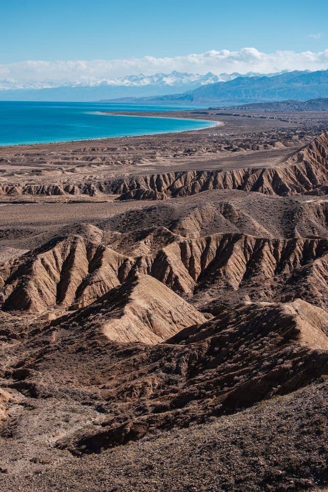 Calanchi bruni e erosi che si affacciano su un grande lago turchese con una catena montuosa innevata in lontananza.