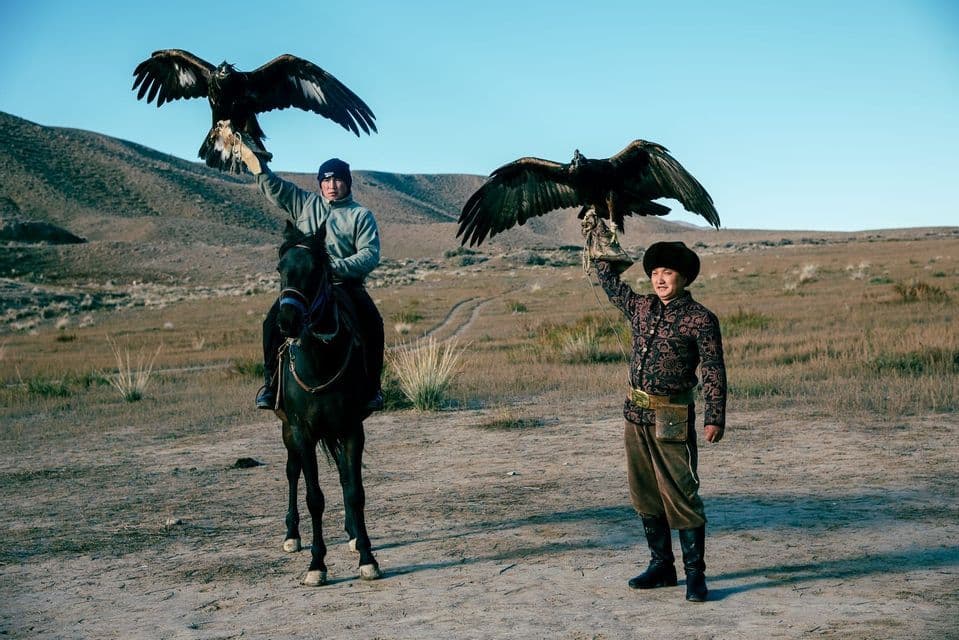 Ein Mann auf einem dunklen Pferd und ein stehender Mann halten zwei Adler mit ausgebreiteten Flügeln in einer kargen, hügeligen Landschaft.