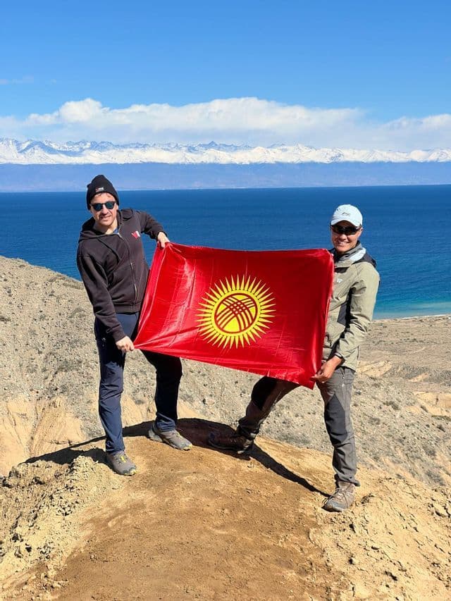 Due uomini di un viaggio di gruppo WeRoad tengono la bandiera del Kirghizistan su una collina che domina un lago e montagne innevate.