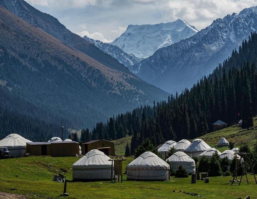 Un villaggio di yurte bianche riposa in una valle verde ai piedi di montagne boscose, con una cima innevata in lontananza.