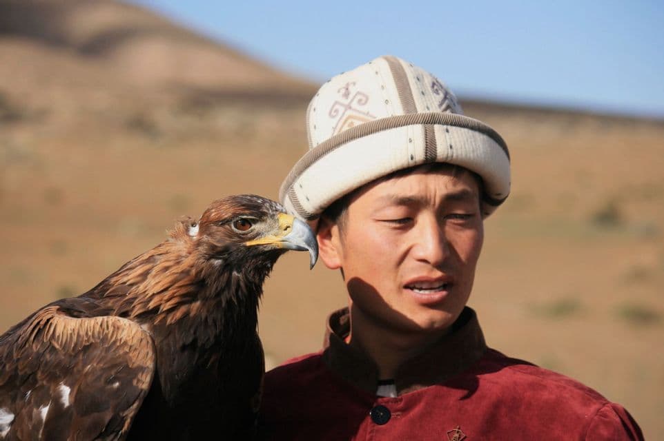 Un uomo con un cappello tradizionale e una giacca rossa si staglia con una grande aquila reale su uno sfondo collinare.