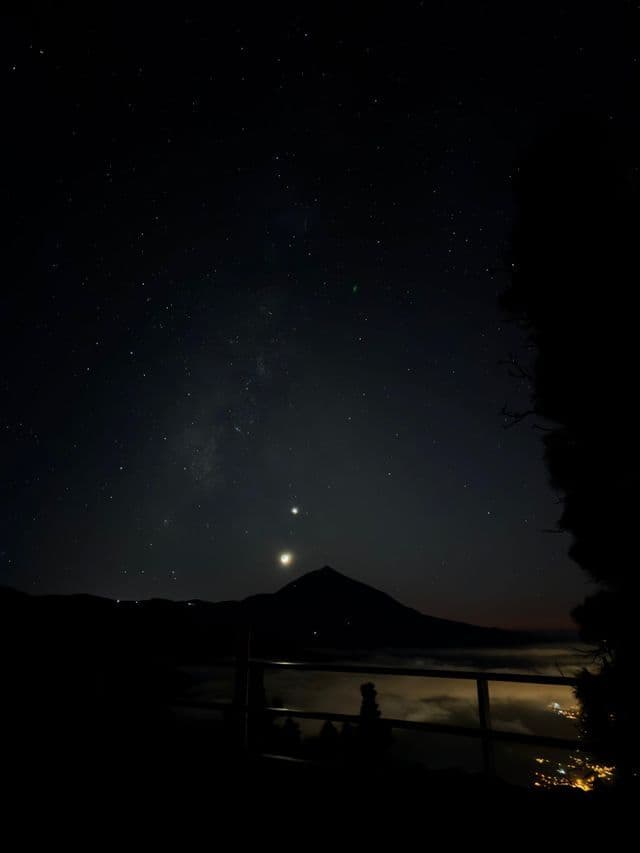 Un cielo notturno stellato con due pianeti luminosi sopra una montagna in silhouette e un mare di nuvole.