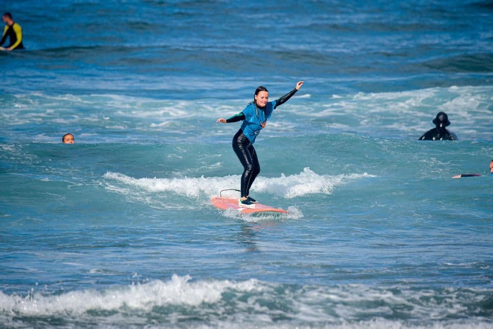 Eine Frau von einer WeRoad Gruppenreise in einem Neoprenanzug steht erfolgreich auf ihrem Surfbrett auf einer blauen Welle.