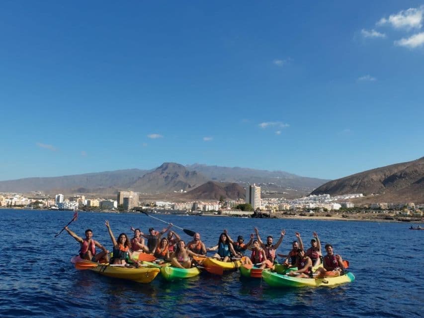 Un gruppo WeRoad in posa su kayak colorati in mare, con una città costiera e montagne sullo sfondo sotto un cielo blu.