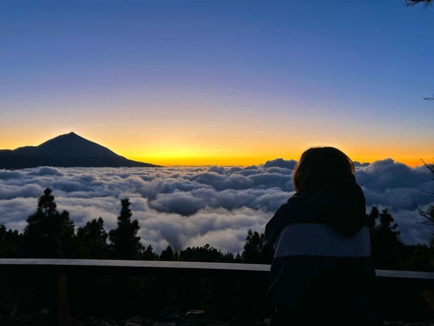 Eine Person, von hinten gesehen, blickt bei Sonnenuntergang auf einen Berggipfel, der sich über ein Wolkenmeer erhebt.