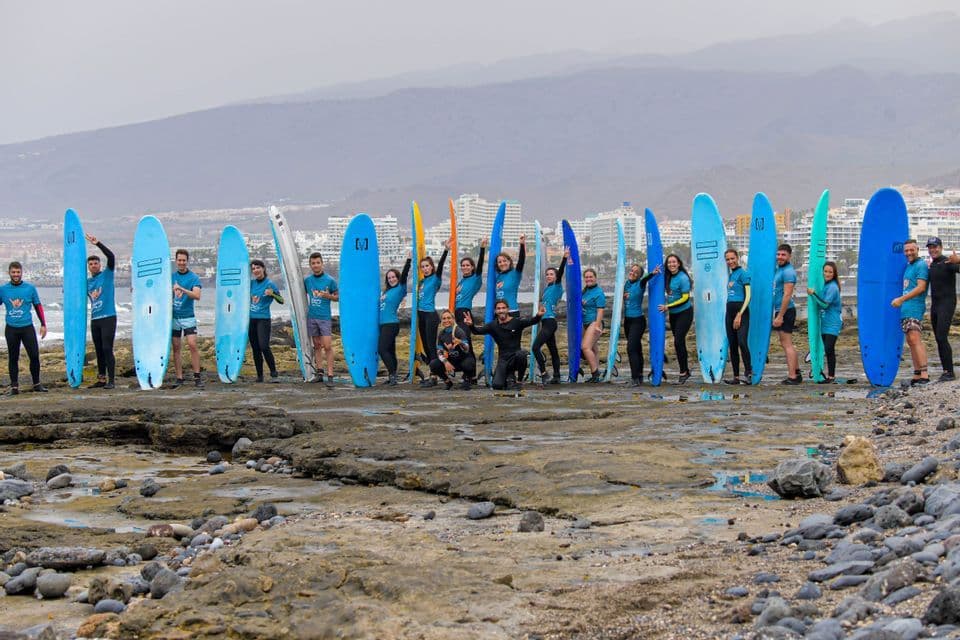 Eine WeRoad-Gruppenreise posiert mit ihren bunten Surfbrettern an einem felsigen Strand vor einer Küstenstadt.