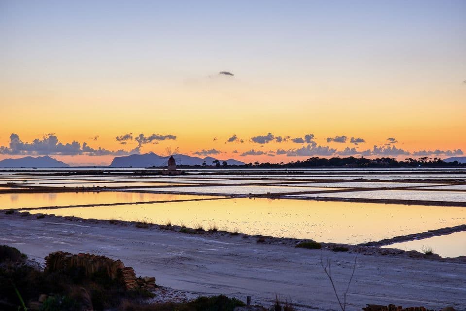 Un molino de viento se alza entre las salinas reflejando un colorido atardecer, con montañas distantes silueteadas contra el cielo naranja.