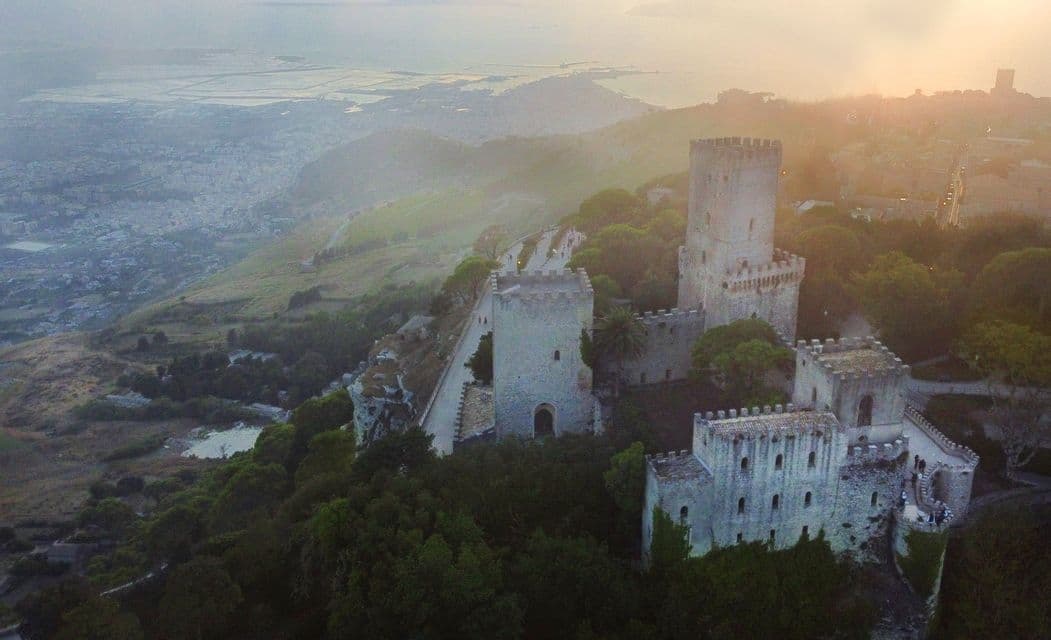 Vista aérea de un gran castillo de piedra con torres en una colina boscosa, con vistas a un valle brumoso y la costa al amanecer.