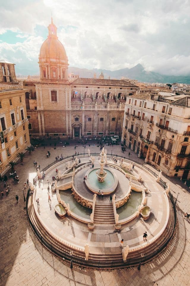 Una vista aérea de una fuente ornamentada y de varios niveles en una plaza histórica, con una cúpula de iglesia iluminada por el sol y montañas al fondo.