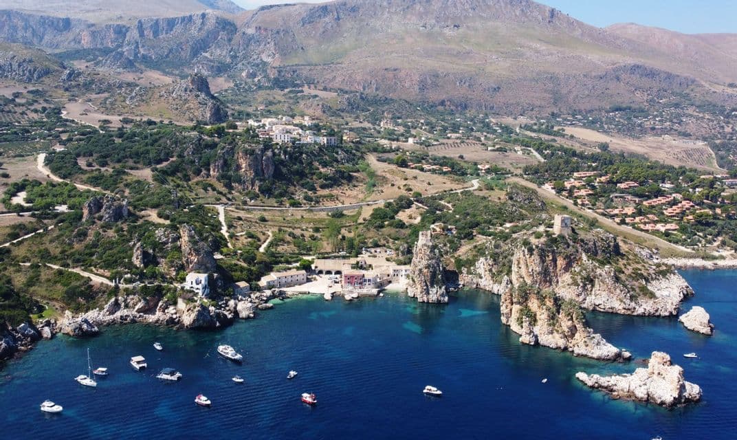 Una vista aérea de un pueblo costero enclavado en una cala rocosa, con varios barcos flotando en el mar azul claro.