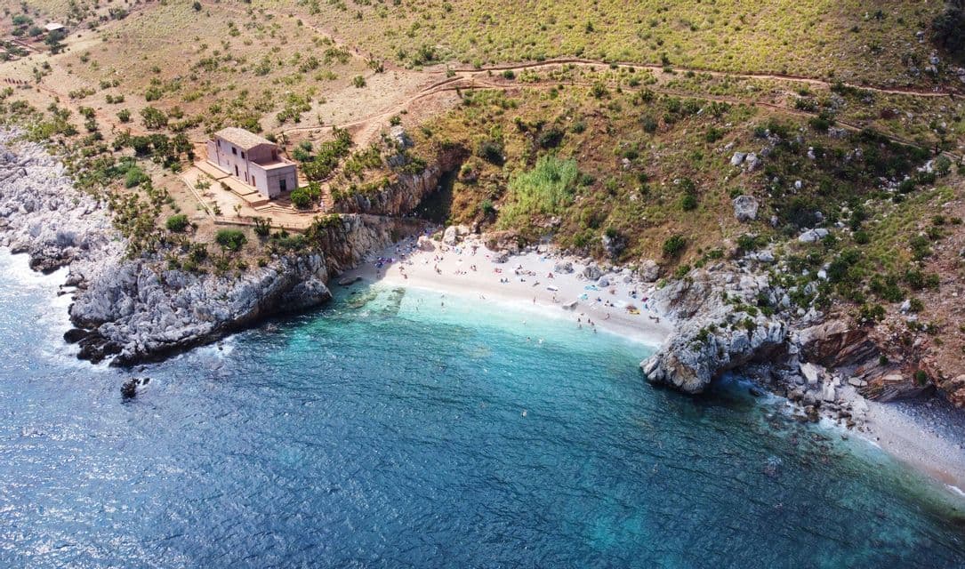 Vista aérea de una cala rocosa aislada con playa de arena, agua turquesa y un edificio de piedra en un acantilado.