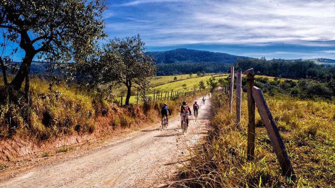 Un gruppo WeRoad percorre in bicicletta un sentiero sterrato in un paesaggio collinare e verdeggiante sotto un cielo azzurro.