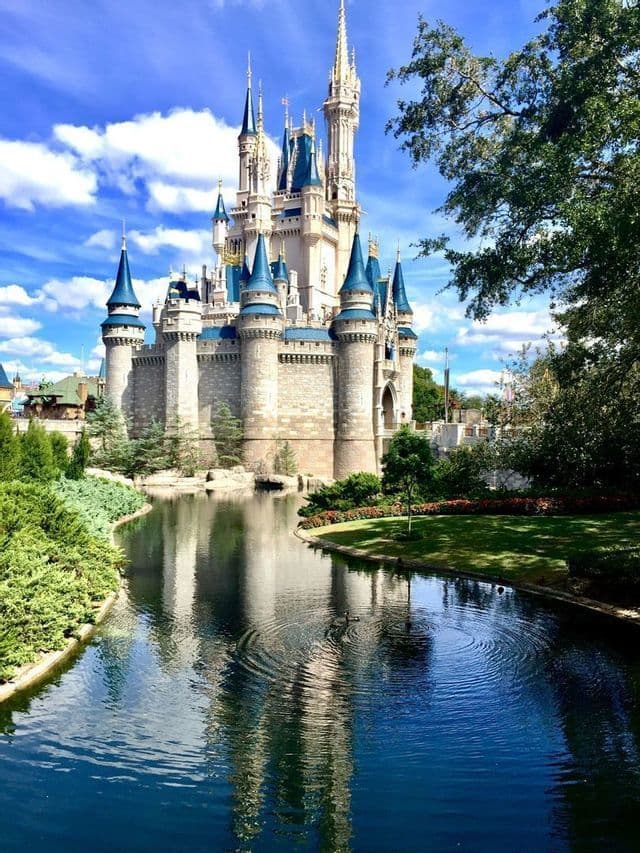 Un gran castillo de piedra con agujas azules se refleja en el agua de un foso circundante en un día soleado con nubes.