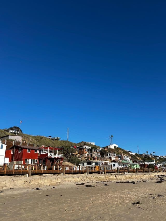 Una hilera de casas coloridas en una ladera cubierta de hierba junto a una playa de arena bajo un cielo azul claro.