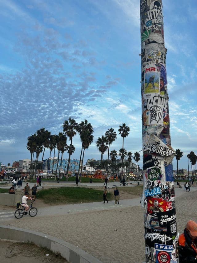 Un poste cubierto de pegatinas en una playa de arena, con gente caminando y en bicicleta cerca de palmeras bajo un cielo nublado.