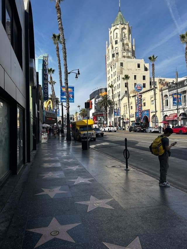 Una persona con una mochila amarilla está de pie en la acera del Paseo de la Fama de Hollywood, bordeada de estrellas y palmeras en un día soleado.