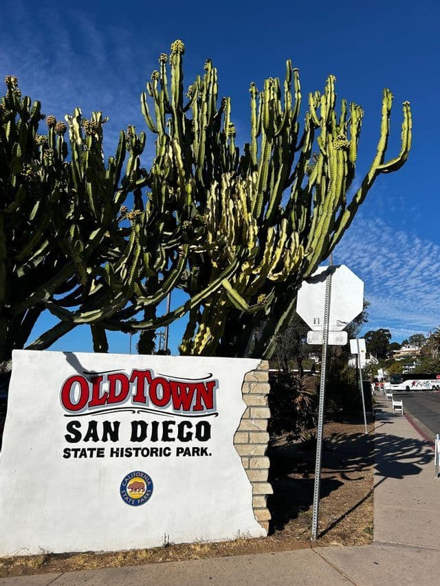 El cartel de bienvenida del Parque Histórico Estatal Old Town San Diego, con un gran cactus detrás sobre un cielo azul claro.
