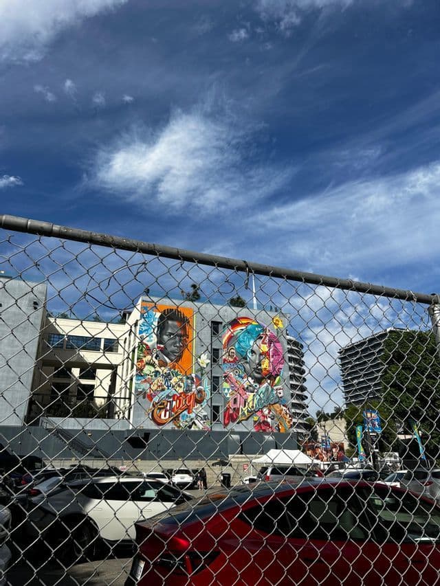 Un gran mural colorido en un edificio se ve a través de una valla de tela metálica, con coches aparcados debajo bajo un cielo azul nublado.