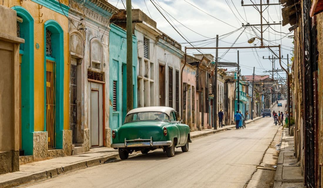 Un coche verde antiguo estacionado en una calle de la ciudad junto a una fila de edificios coloridos y desgastados bajo un cielo nublado.