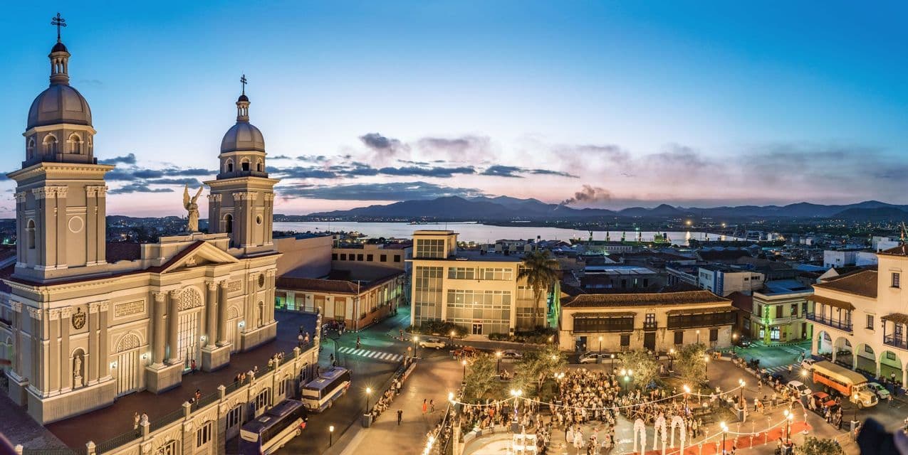 Una vista panorámica elevada de una plaza de la ciudad y una catedral al atardecer, con una bahía y montañas visibles a lo lejos.