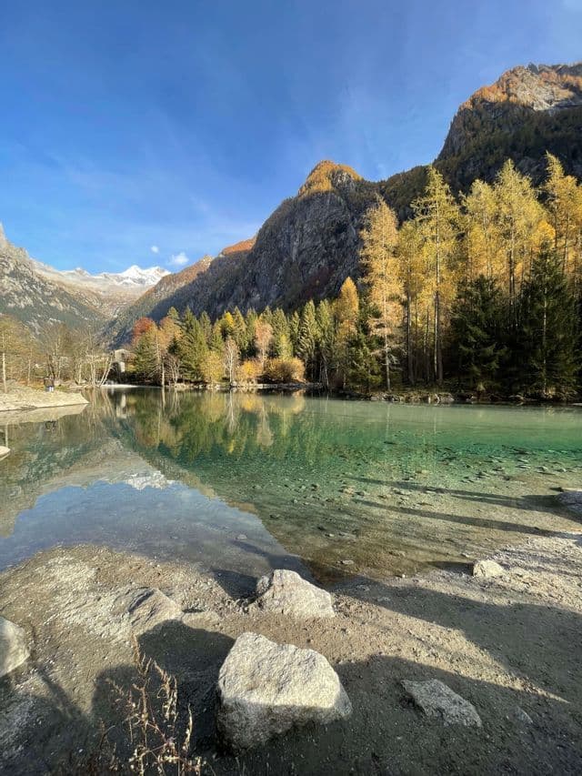 Una sponda rocciosa conduce a un limpido lago alpino che riflette una foresta autunnale e montagne innevate sotto un cielo blu.
